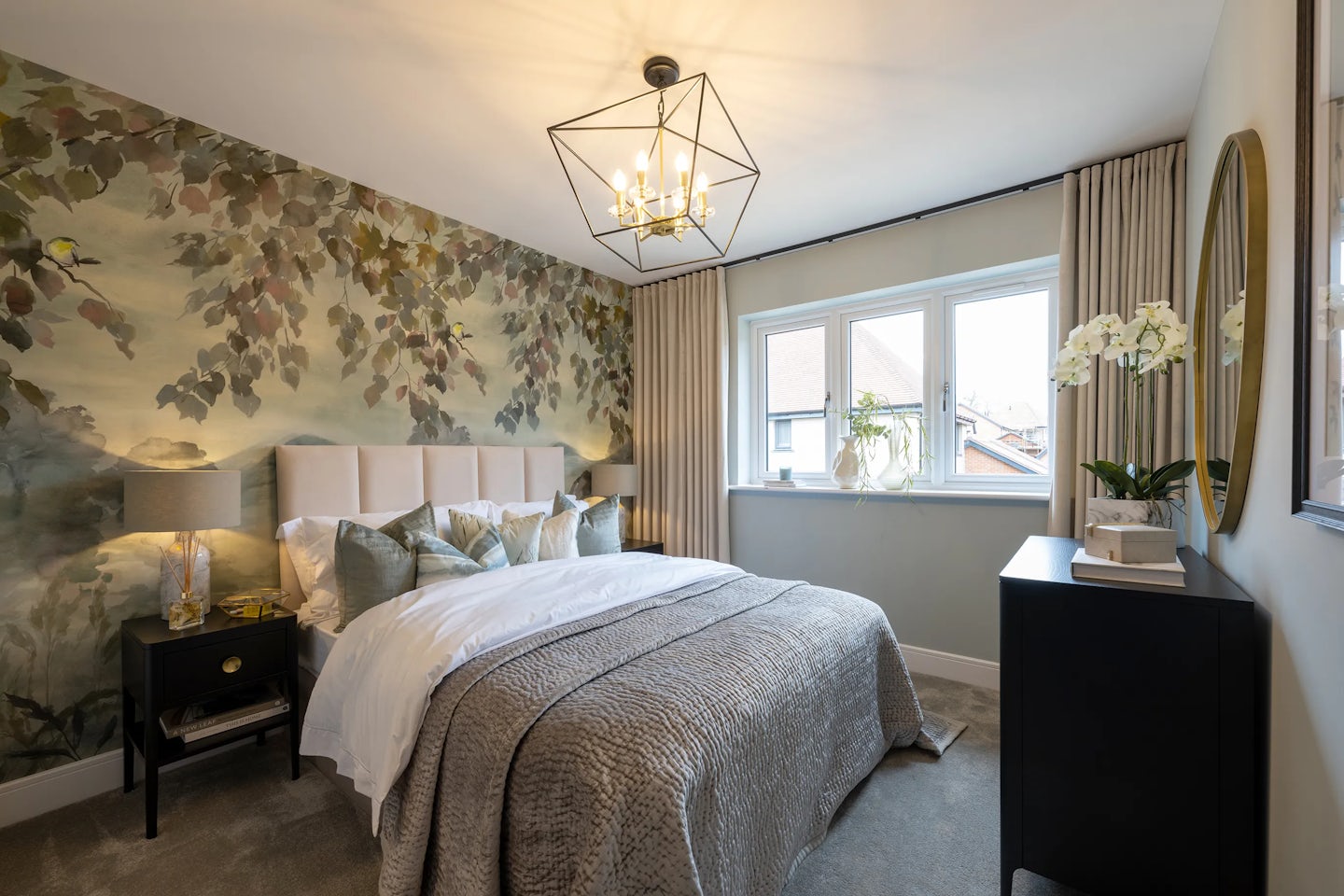 Well-appointed bedroom featuring a double bed with a beige upholstered headboard and layers of white, green, and patterned pillows atop a white duvet and textured grey blanket. A nature-inspired mural with leafy branches spans the accent wall behind the bed, complemented by a geometric chandelier with exposed bulbs. On the left, a black nightstand holds a lamp and decorative items; on the right, a matching dresser supports a white orchid and neatly arranged books.