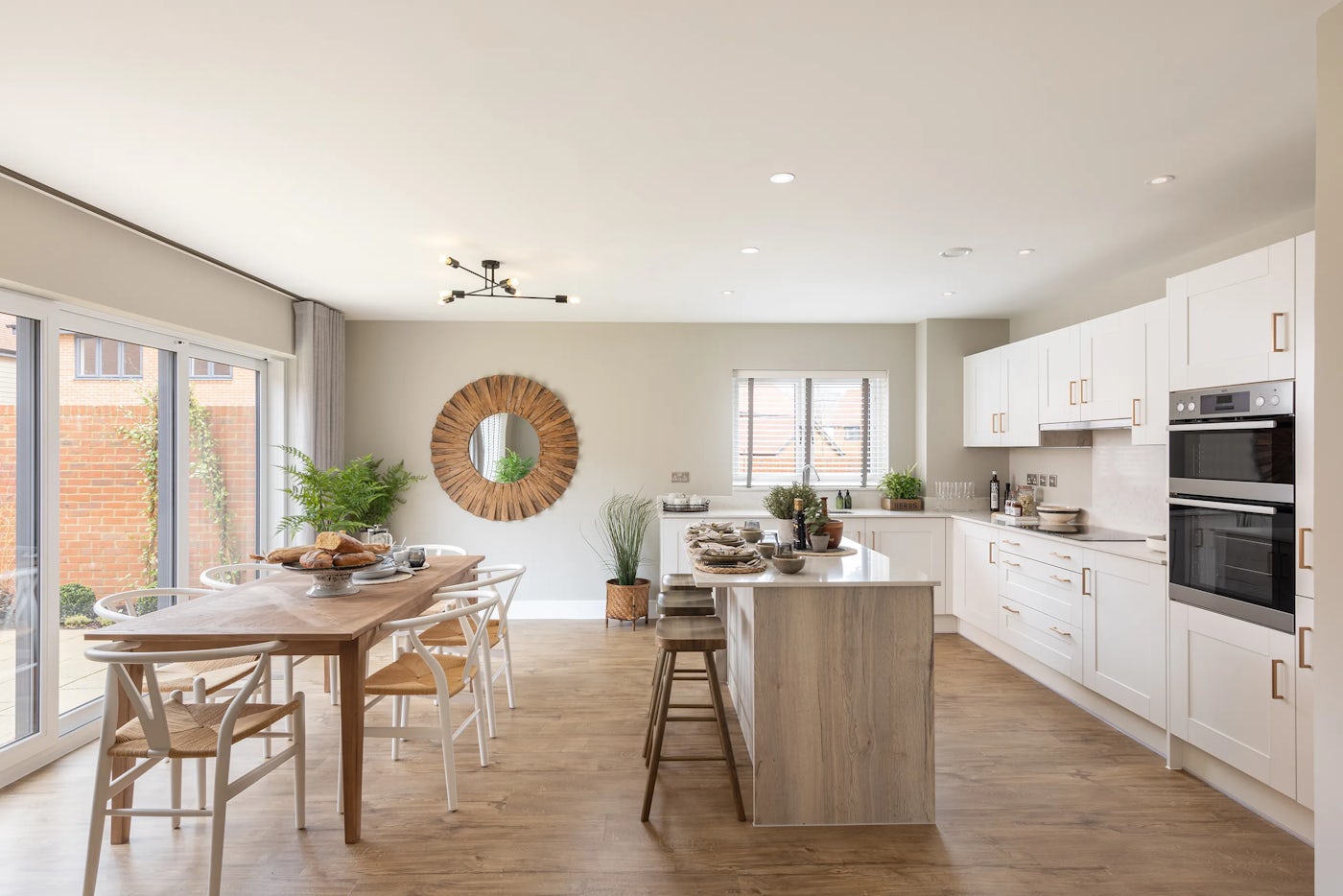 Contemporary kitchen and dining area styled in soft neutrals, featuring white cabinetry with gold handles and built-in appliances including an oven and microwave. A central island with a light wood finish includes bar stools for casual seating. Adjacent is a wooden dining table surrounded by white chairs, topped with baskets of bread, croissants, and fruit for a warm, inviting touch. Large glass doors lead to an outdoor space with a brick wall and lush greenery
