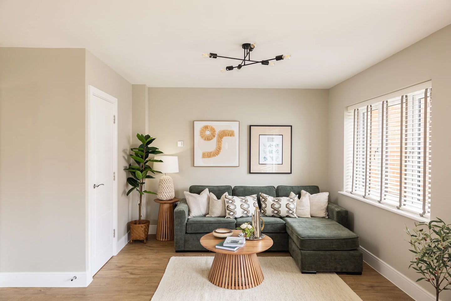 Modern living room with a green sectional sofa styled with patterned throw pillows, positioned beside a round wooden coffee table holding books and decorative objects. A light rug anchors the seating area, while two abstract artworks hang above the sofa on a beige wall. A floor lamp and potted plant sit to the side, adding warmth and texture. Natural light streams in through a large window with white blinds