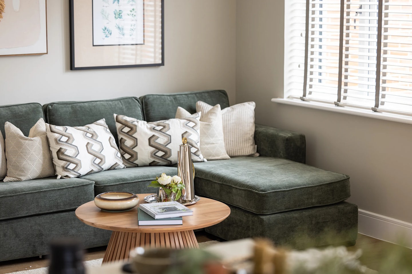 Inviting living room with a green sectional sofa adorned with patterned cushions, centered around a round wooden coffee table holding a decorative tray, vase with flowers, books, and a bowl. Natural light streams through white blinds covering a large window, illuminating beige walls and wood flooring. Above the sofa, two framed abstract artworks add visual interest.
