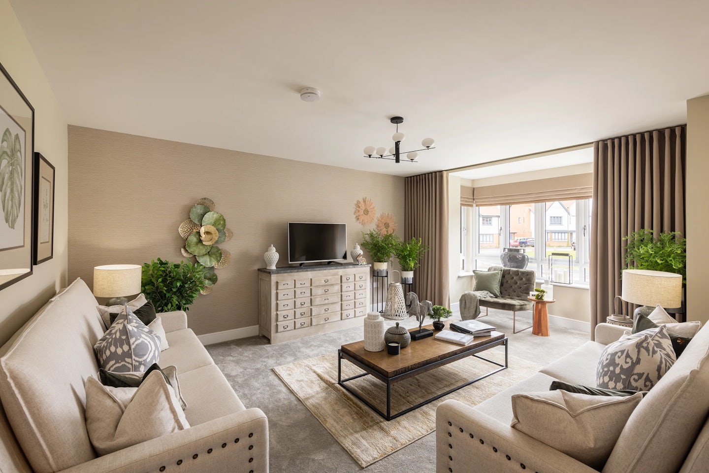 Warm, inviting living room with two beige sofas adorned with patterned cushions, arranged around a wooden coffee table styled with books, candles, and a small plant. A large textured rug anchors the space, while a wooden sideboard supports a television and additional decor including framed art and greenery. Table lamps sit atop matching side tables, casting a gentle glow. Soft natural light filters through a wide window dressed with blinds and curtains, and a geometric chandelier adds a contemporary touch overhead.