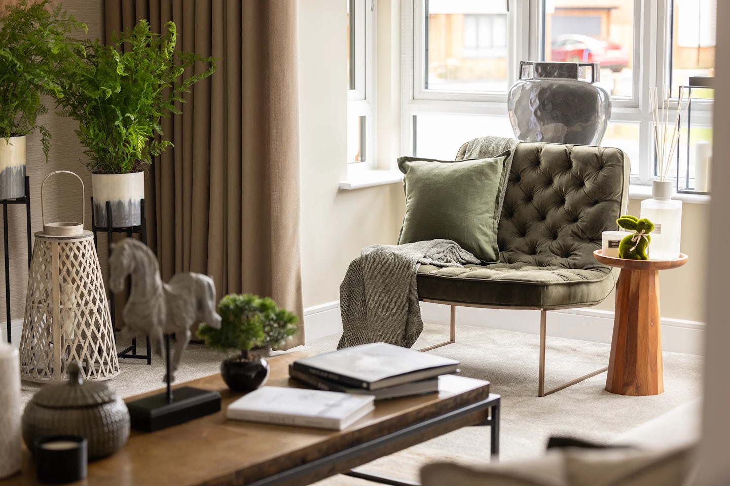 Cozy living room corner featuring a tufted green armchair with a green cushion and gray throw, set beside a wooden side table topped with a potted plant. A large window behind the chair filters soft daylight, while the surrounding decor includes tall leafy plants, a lantern, and a coffee table styled with a bonsai tree, books, and a horse figurine.