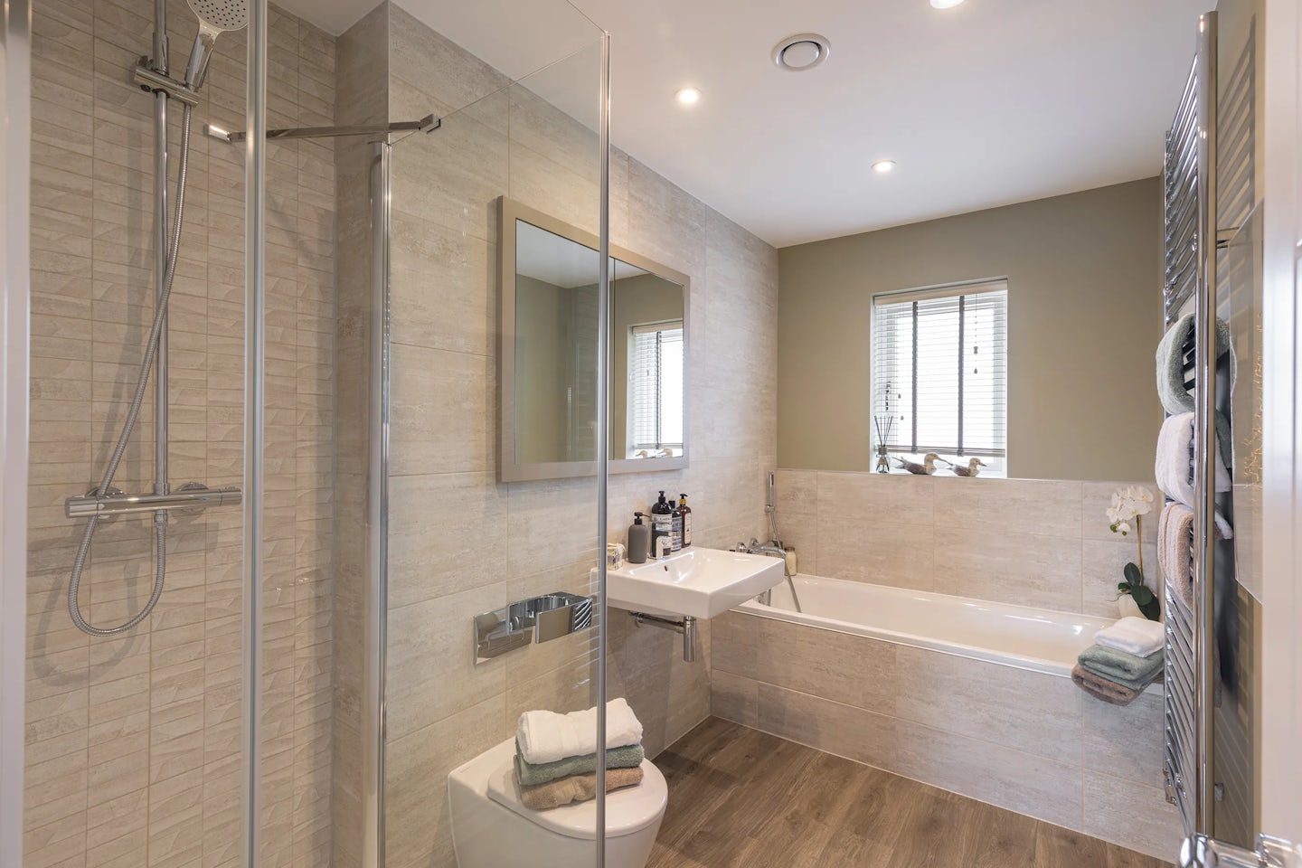Modern bathroom featuring a glass-enclosed shower with chrome fittings, set beside a white toilet topped with neatly folded towels. A large wall-mounted mirror reflects natural light from a nearby window with blinds, enhancing the clean, minimalist space. To the right, a white sink holds assorted toiletries, and a rectangular bathtub is styled with a green plant and a hanging towel.