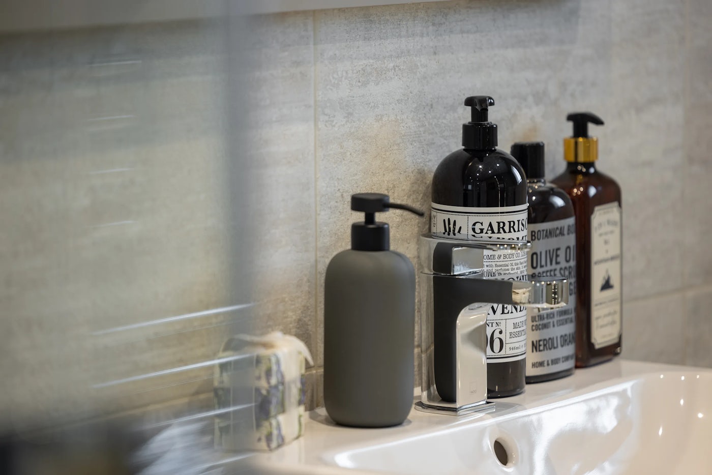 Bathroom sink area featuring a modern metallic faucet mounted on a white countertop, surrounded by neatly arranged toiletries. Items include a grey pump bottle, a black bottle labeled “GARRISON,” a transparent bottle with a dark label, and a brown bottle labeled “BOTANICAL APOTHECARY.” A bar of soap sits to the left of the faucet, while beige tiles form a clean, neutral backdrop.