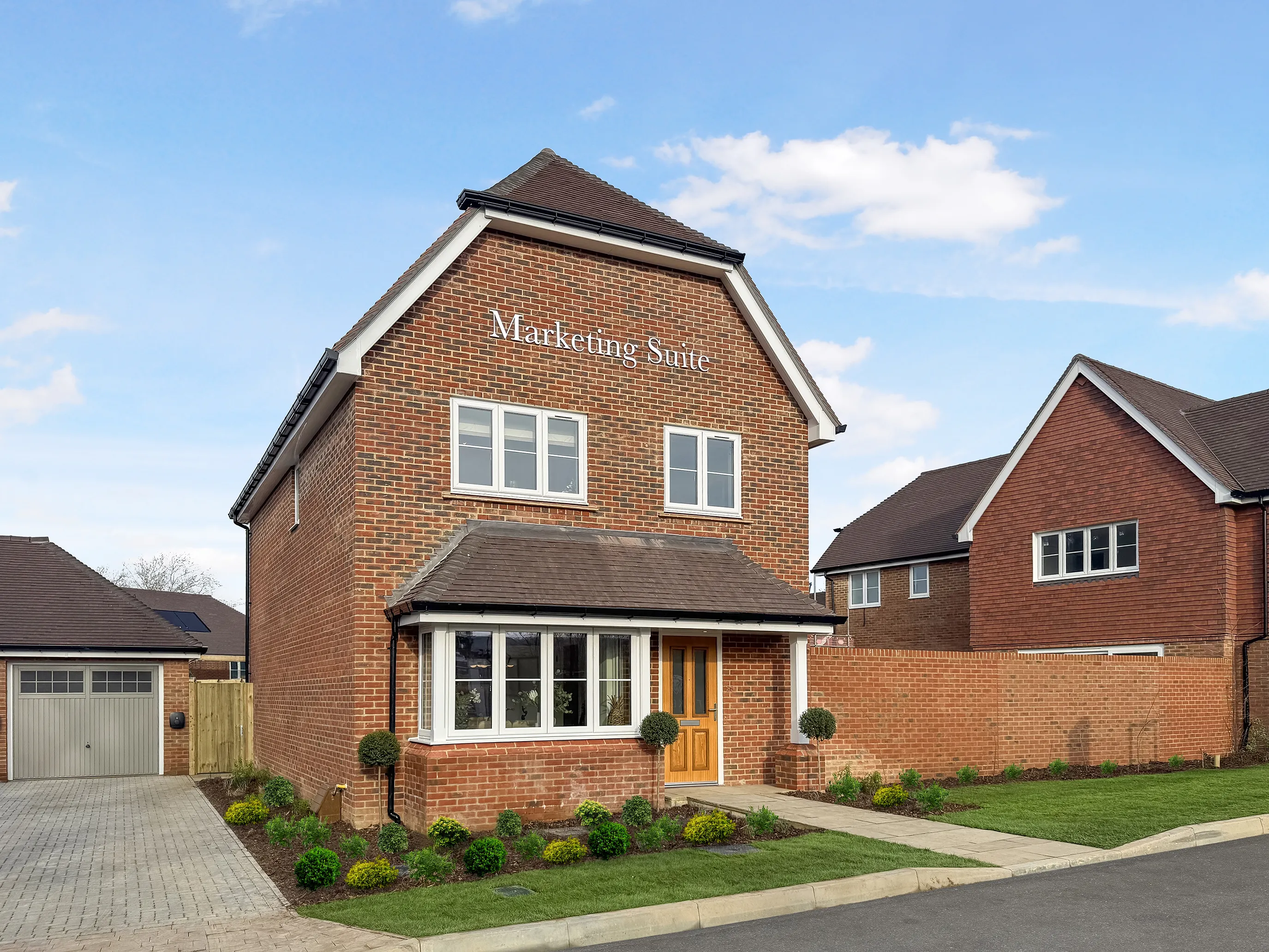 Modern two-story brick house featuring a pitched roof with dark shingles and white-trimmed windows. A wooden front door is set beneath a covered porch, flanked by neatly trimmed hedges and a small front lawn. A paved driveway leads to a garage on the left, while a similar neighboring house appears in the background.
