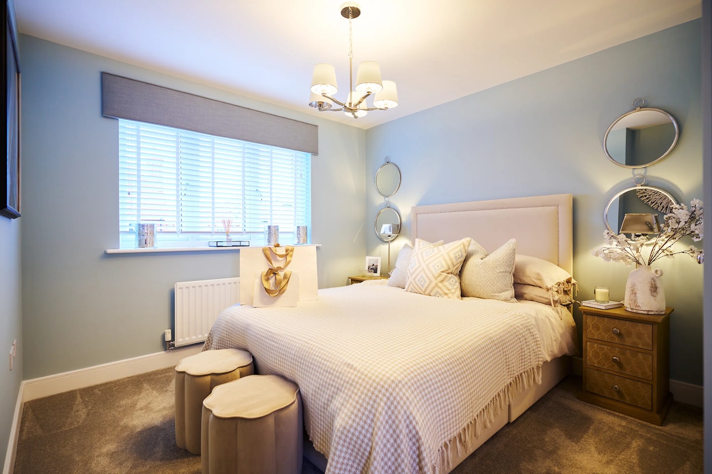 Tranquil bedroom featuring a large bed with a beige upholstered headboard and checkered bedspread, accented by layered pillows in geometric patterns. Three beige ottomans sit at the foot of the bed atop a soft brown carpet. To the right, a wooden nightstand with three drawers holds a lamp, a vase of white flowers, and decor, framed above by two round mirrors. A wide window with white blinds and a grey valance allows natural light in, highlighting candles and small vases on the windowsill. Pale blue walls and a three-shade chandelier overhead complete the cozy, polished atmosphere