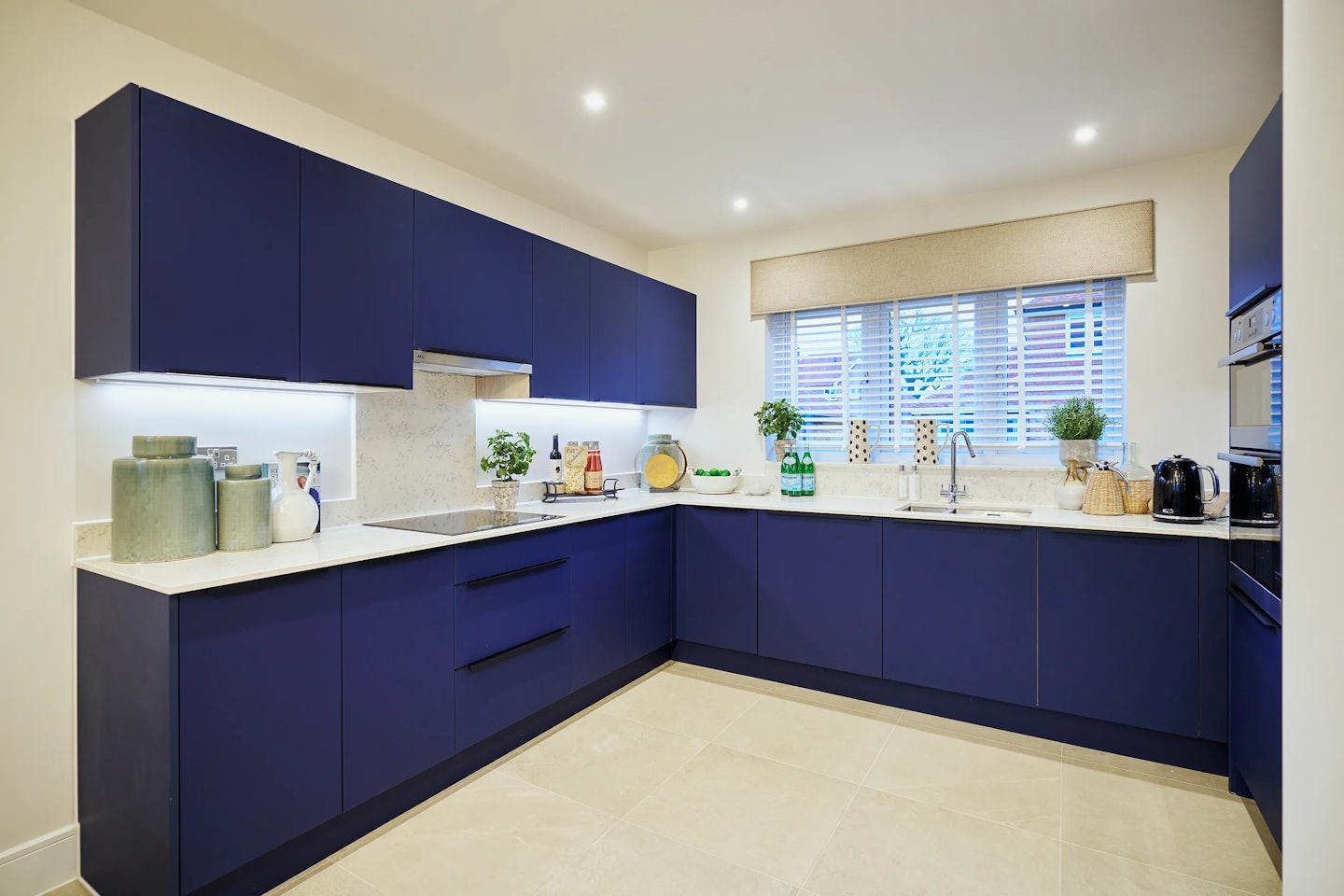 Contemporary U-shaped kitchen with deep blue cabinetry and sleek white countertops, set against warm beige tiled flooring. Natural light filters through a window dressed with blinds and a matching valance, illuminating details like green glass jars, a potted plant, a fruit bowl, and a wooden cutting board with oil and seasoning. A white vase and a chrome kettle add functional elegance, while recessed ceiling lights enhance the clean, inviting feel of the space