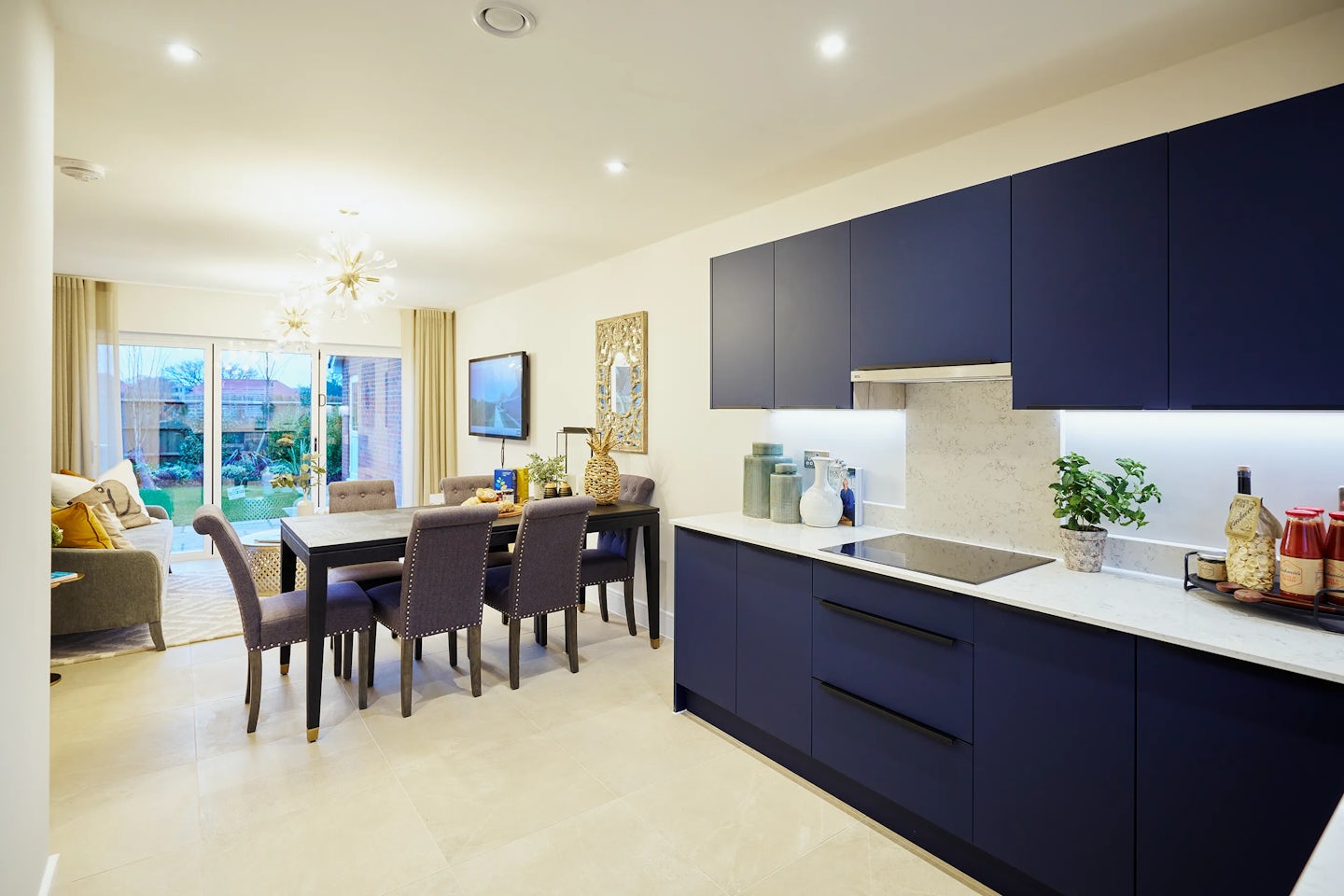 Open-plan interior combining a modern kitchen, dining area, and living space. The kitchen features deep blue cabinetry, white countertops, and an induction cooktop, complemented by decorative vases and potted plants. The adjacent dining area includes a rectangular table surrounded by six upholstered chairs beneath a glass pendant chandelier, with a large wall-mounted mirror enhancing depth. In the background, a neutral-toned sofa sits opposite sliding glass doors that lead to an outdoor area, allowing natural light to brighten the cohesive, stylish layout