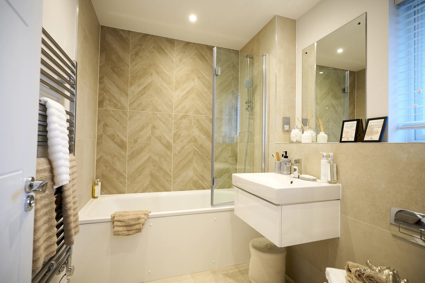 Modern bathroom featuring a herringbone-tiled wall in beige tones behind a white bathtub with a glass shower screen and chrome showerhead. To the right, a rectangular mirror hangs above a white sink styled with assorted toiletries. Below are built-in cabinets offering clean-lined storage. Two framed certificates or artworks are mounted beside the mirror, adding a personal touch. A towel rack with neatly folded white towels sits to the left, and recessed ceiling lights provide soft, even illumination throughout the space