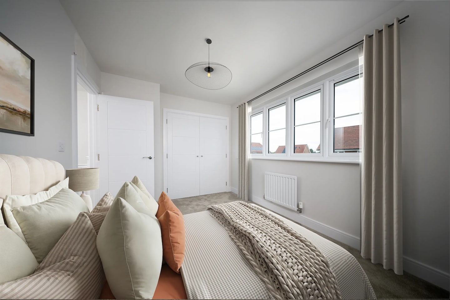 Contemporary bedroom featuring a double bed with a beige upholstered headboard, dressed in layered pillows in muted tones of green, orange, and beige. A knitted blanket adds texture atop the crisp white bedding. To the left, a bedside table supports a sleek lamp, and a framed painting hangs on the adjacent wall. Light streams through a large window with white frames and beige curtains, illuminating soft carpeted flooring and pale walls. A radiator sits below the window, and two white doors on the far wall suggest access to storage or a separate room. Overhead, a modern light fixture enhances the cozy, composed atmosphere