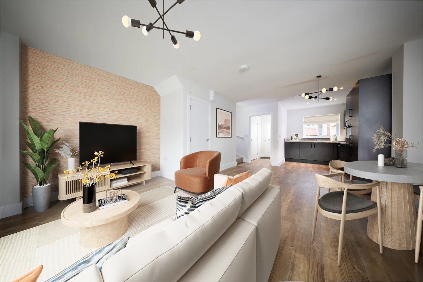 Contemporary open-plan living space combining a lounge, dining area, and partial kitchen view. The lounge features a large flat-screen TV atop a wooden console flanked by a potted plant and a vase of yellow flowers. Seating includes two brown armchairs and a white sofa with patterned cushions, arranged around a circular wooden coffee table holding books and decor. Light wood flooring and a textured beige accent wall add warmth. In the background, dark kitchen cabinets and a pale countertop complement a small round dining table with two wooden chairs. Multiple pendant bulbs illuminate the cohesive, stylish atmosphere