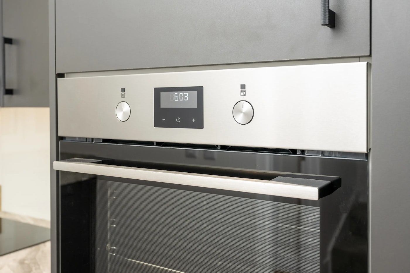 Close-up of a built-in kitchen oven with a stainless steel control panel featuring two knobs and a central digital display reading “6:03.” The oven door includes a glass window and a horizontal handle, offering a partial glimpse into the interior. The unit is set seamlessly into a light-toned cabinet, with a portion of the countertop visible on the left, contributing to a modern, integrated kitchen aesthetic