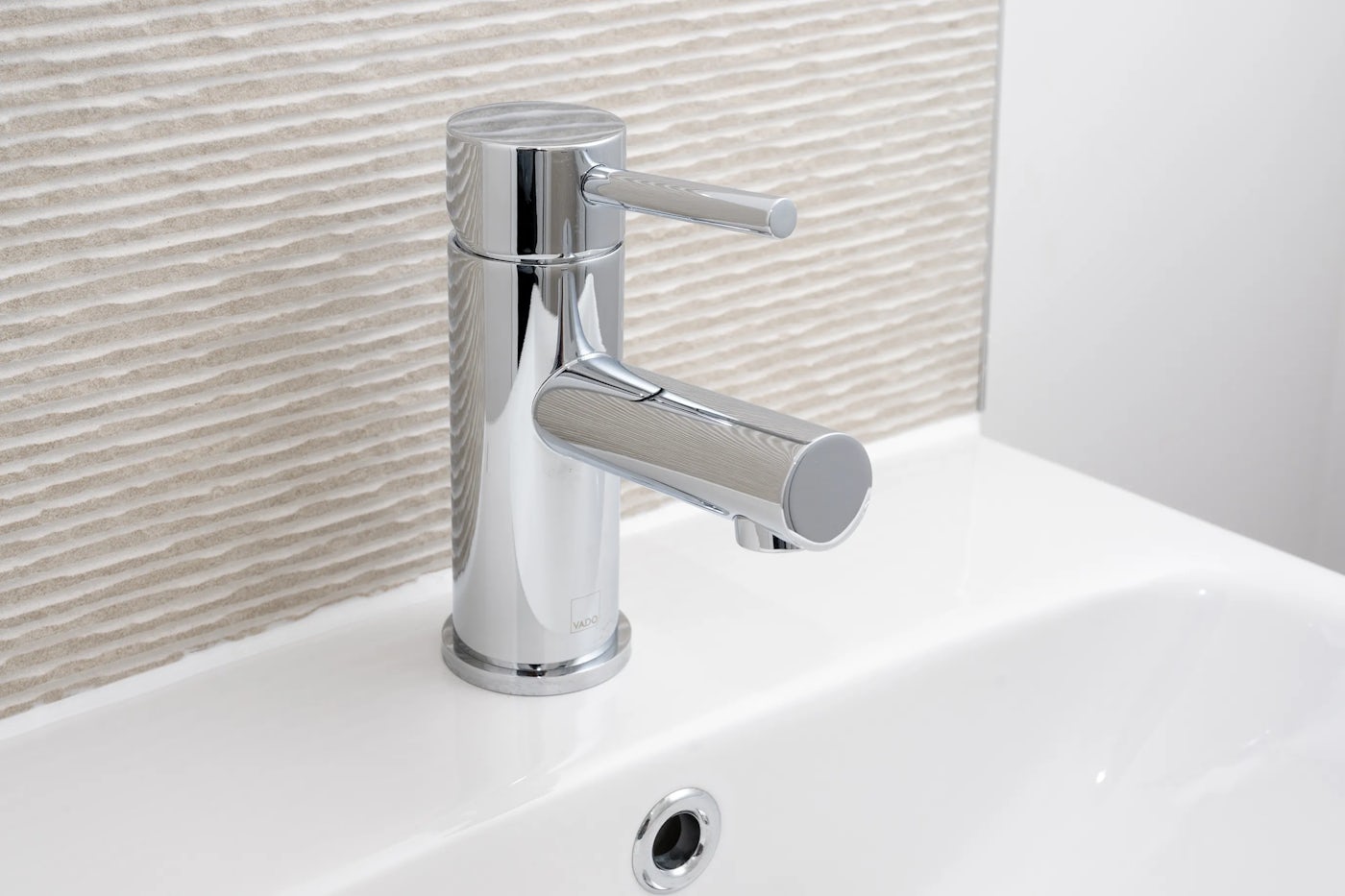 Close-up of a chrome-finished faucet with a cylindrical base and single lever handle, mounted on a white basin. The backdrop features a textured, beige wall that adds warmth to the minimalist aesthetic. The faucet’s reflective surface and clean lines convey a sleek, modern design suited to a contemporary bathroom or kitchen setting