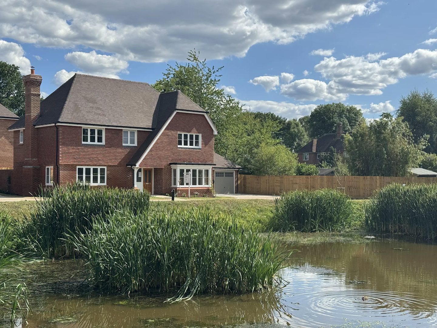 Idyllic countryside scene featuring a large two-story red brick house with white-framed windows, a chimney, and a pitched tiled roof. The home is situated behind a tall wooden fence, bordered by a lush pond in the foreground, where green reeds and grasses flourish along the water’s edge. Trees and additional houses peek through the greenery in the background, under a partly cloudy blue sky that bathes the landscape in soft light. The setting blends natural beauty with rural residential charm.
