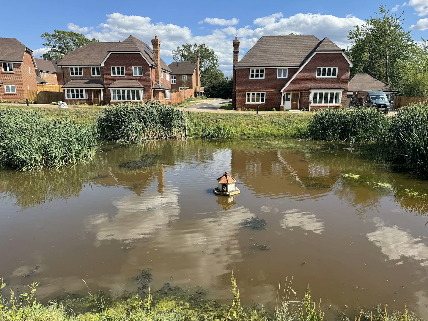 Small pond set within a tranquil residential green space, centered by a wooden birdhouse-like structure on stilts above the water. The pond is bordered by tall grasses and reeds, with reflections of scattered clouds visible on its calm surface. In the background, red-brick houses with pitched roofs and prominent chimneys line the horizon, partially framed by leafy trees. The blue sky and soft sunlight enhance the peaceful, natural charm of the suburban setting.