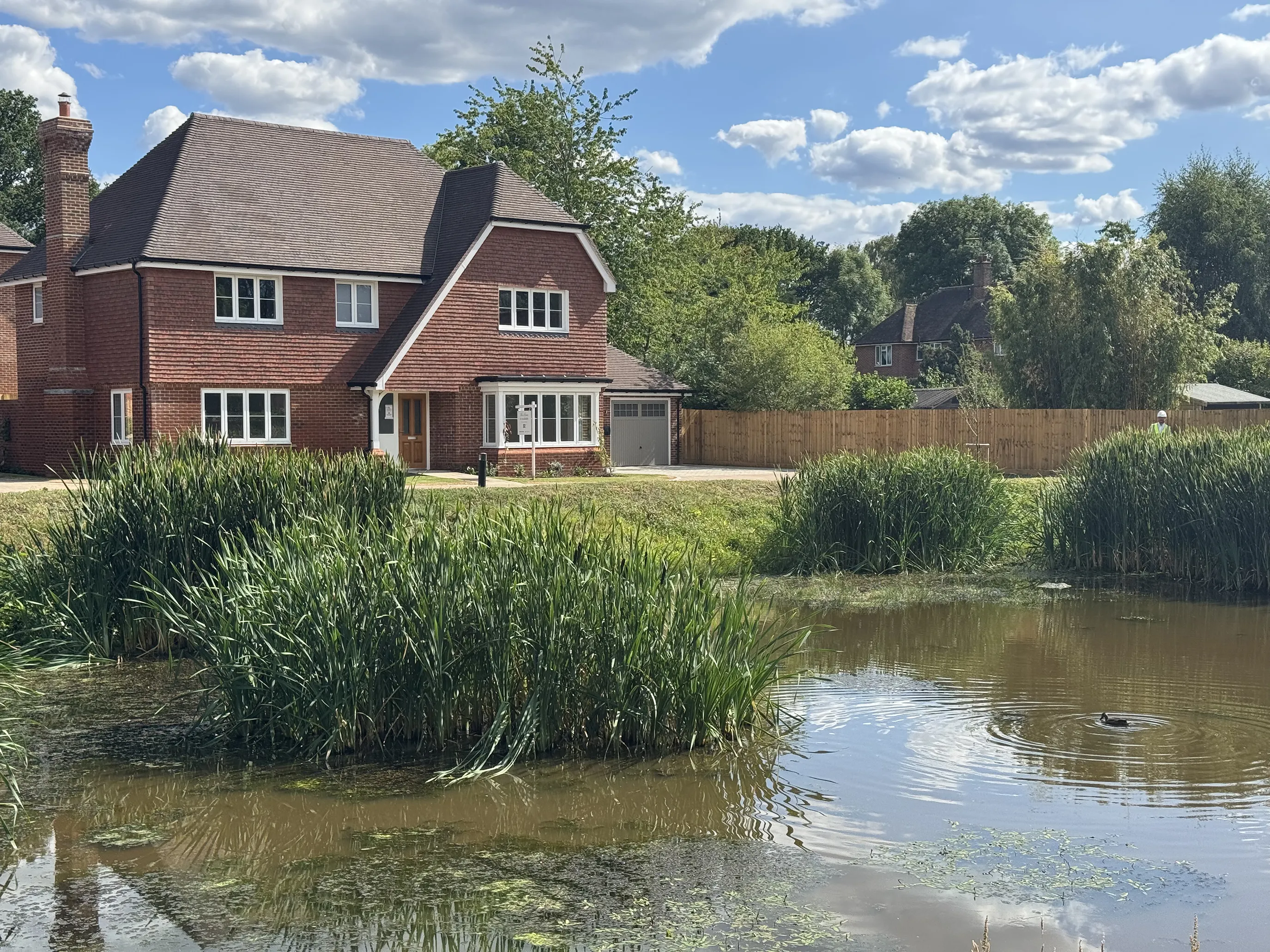 Picturesque countryside home with a two-story red brick exterior and brown tiled roof, surrounded by neatly landscaped greenery. The house features multiple white-framed windows and a small porch over the front door. In the foreground, a tranquil pond with clear water is bordered by tall green reeds and flowering aquatic plants, separated from the garden by a wooden fence. The bright blue sky with scattered clouds enhances the peaceful, rural charm of the setting.