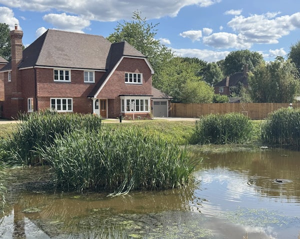 Picturesque countryside home with a two-story red brick exterior and brown tiled roof, surrounded by neatly landscaped greenery. The house features multiple white-framed windows and a small porch over the front door. In the foreground, a tranquil pond with clear water is bordered by tall green reeds and flowering aquatic plants, separated from the garden by a wooden fence. The bright blue sky with scattered clouds enhances the peaceful, rural charm of the setting.