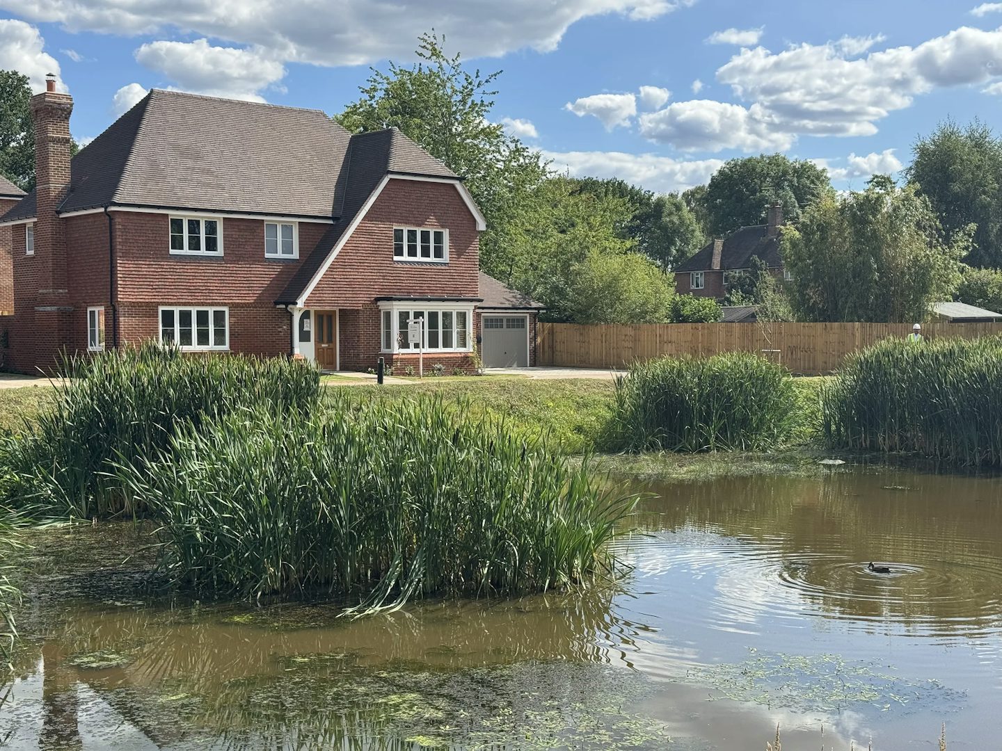 Picturesque countryside home with a two-story red brick exterior and brown tiled roof, surrounded by neatly landscaped greenery. The house features multiple white-framed windows and a small porch over the front door. In the foreground, a tranquil pond with clear water is bordered by tall green reeds and flowering aquatic plants, separated from the garden by a wooden fence. The bright blue sky with scattered clouds enhances the peaceful, rural charm of the setting.
