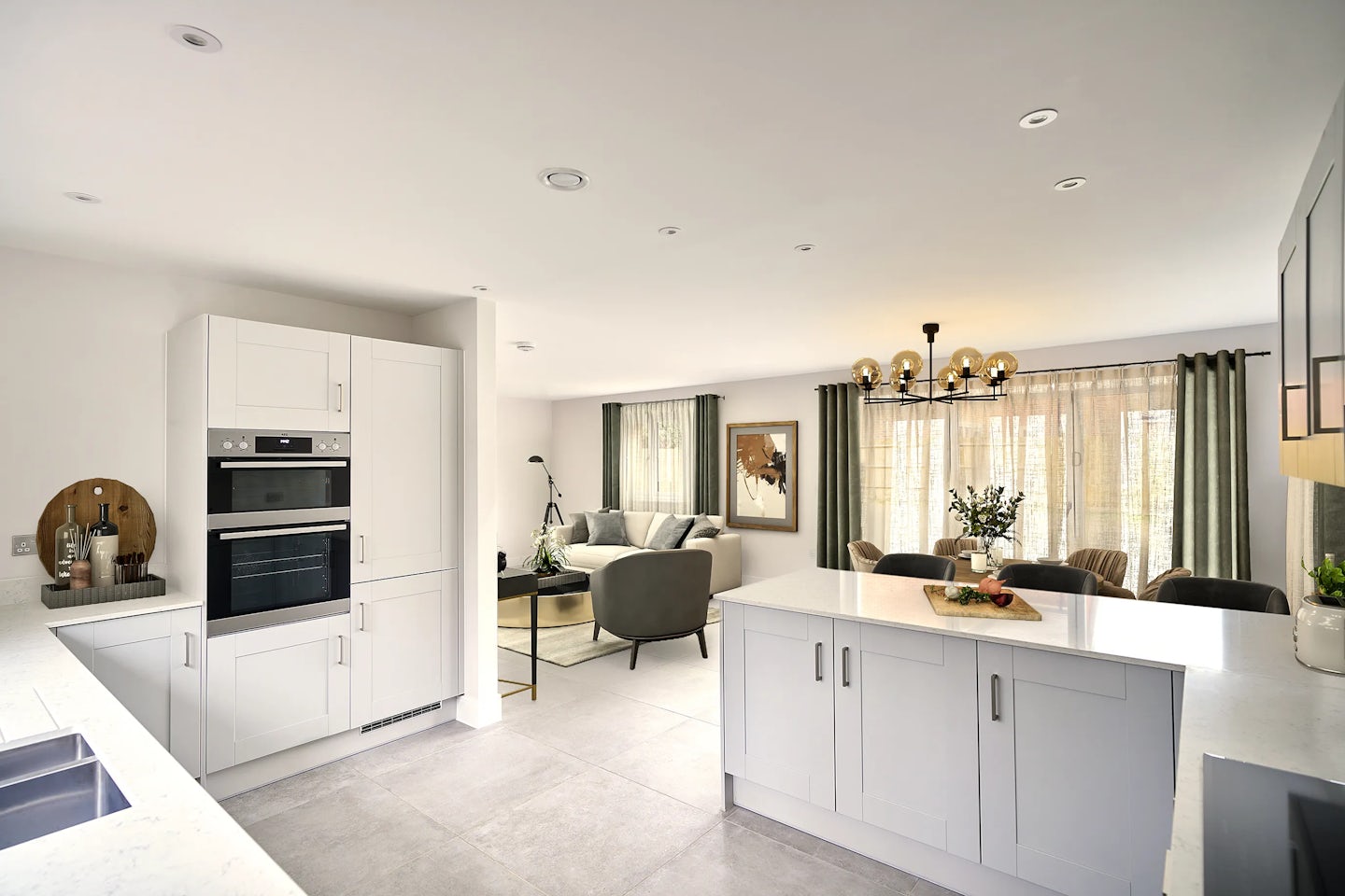 Spacious open-plan kitchen and living area with white cabinetry and built-in appliances, including a microwave and oven. A central island with a waterfall-edge white countertop features a chopping board with fresh vegetables and a knife, suggesting meal prep. In the background, the dining space includes a wooden table with a vase of flowers and a chandelier composed of multiple exposed bulbs. The living area offers a sofa, cushioned chair, round mirror, and wall art, all illuminated by soft natural light streaming through large curtained windows.