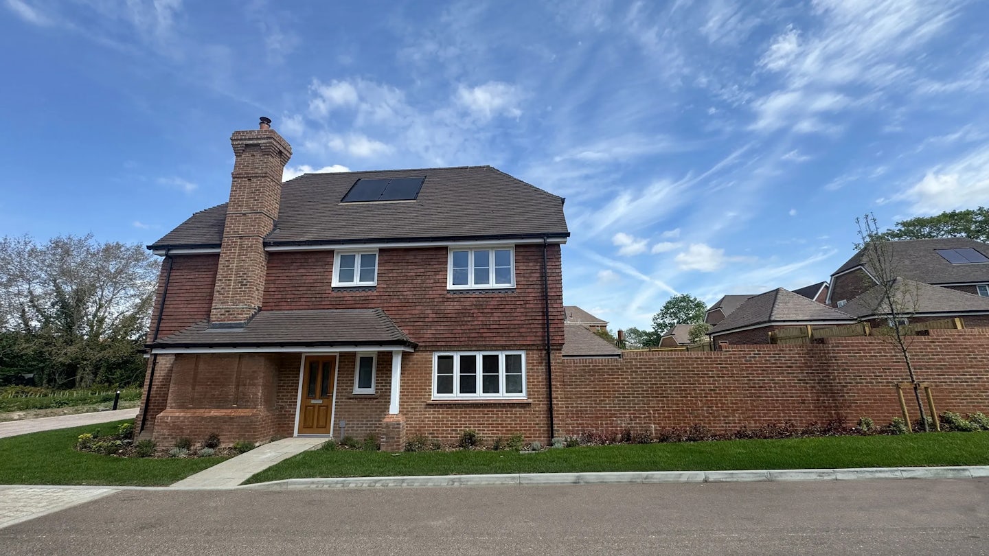 Two-story red brick house with a sloped tiled roof, featuring solar panels and a skylight. A chimney rises from the left side, and a small porch with white framing shelters the central front door, which is flanked by a side window. A neatly maintained lawn and a paved driveway lead up to the home, bordered by a low brick wall enclosing a garden area.