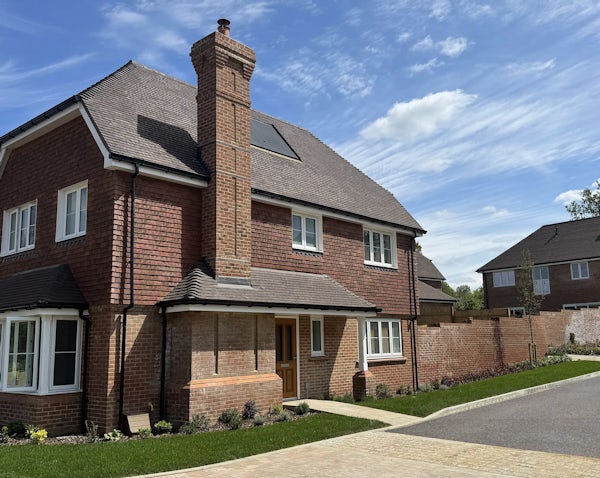 Modern two-story red brick house with a pitched tiled roof and prominent chimney, set within a neatly landscaped front garden and paved driveway. Large white-framed windows line both floors, and a small porch shelters the front door. The home is bathed in bright daylight under a partly cloudy blue sky, while a similar neighboring house is visible in the background