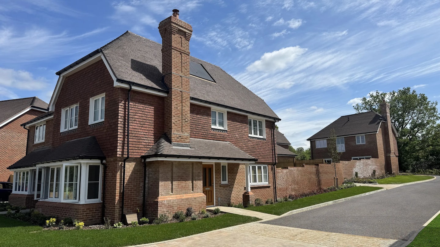 Modern two-story red brick house with a pitched tiled roof and prominent chimney, set within a neatly landscaped front garden and paved driveway. Large white-framed windows line both floors, and a small porch shelters the front door. The home is bathed in bright daylight under a partly cloudy blue sky, while a similar neighboring house is visible in the background