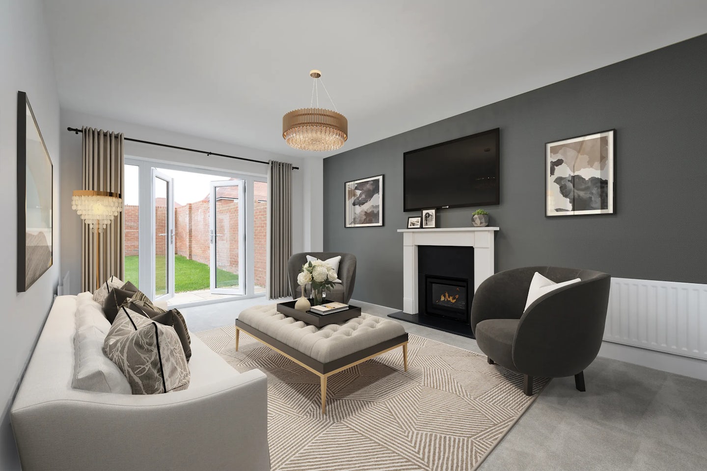 Elegant living room with a soft grey sofa and patterned cushions on the left, facing a large square ottoman topped with a tray and floral arrangement. To the right, a charcoal armchair adds contrast, while a fireplace with a white mantel and black insert anchors the far wall below a mounted flat-screen TV. Flanking the TV are two framed abstract artworks, set against a deep grey accent wall. Natural light streams in through wide glass doors overlooking a garden with a brick wall, and a chandelier adds ambient warmth from above. The light-toned rug and neutral color palette create a sophisticated, serene atmosphere