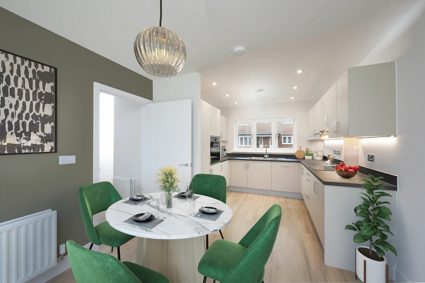 Stylish kitchen and dining area with white cabinetry, black countertops, and stainless steel appliances. A window above the sink lets in natural light, complementing the olive green accent wall and adjacent white surfaces. The dining space features a round marble table set for three, with green upholstered chairs and a vase of yellow flowers as the centerpiece. Recessed ceiling lights and a pendant lamp provide layered illumination, while a potted plant and abstract wall art introduce warmth and personality to the cohesive, contemporary setting