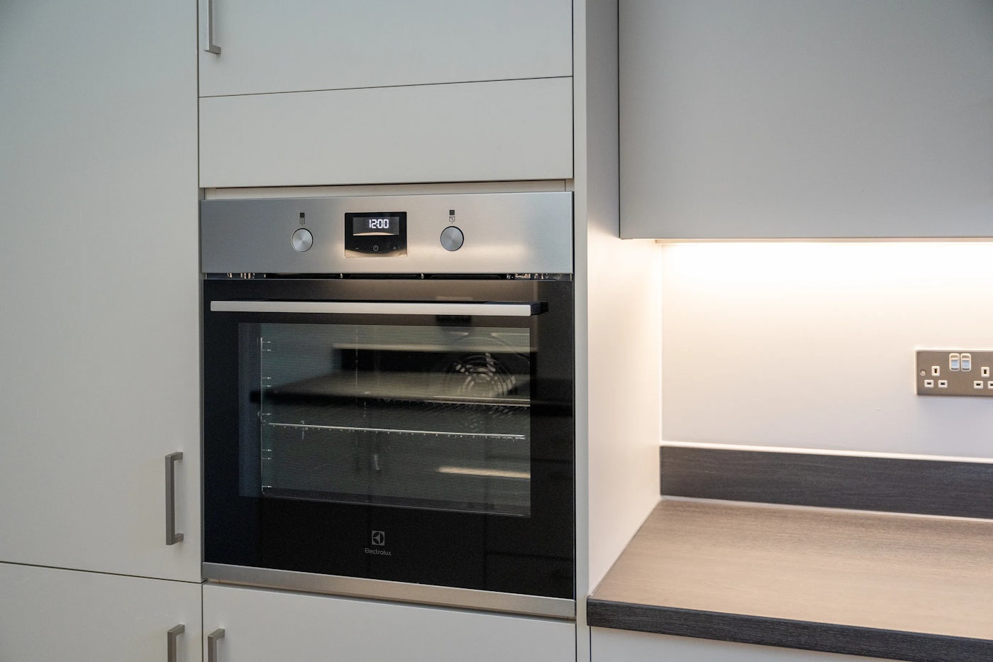 Modern kitchen featuring a built-in oven with a digital display set to “12:00” and two chrome-finished control knobs. The oven is seamlessly integrated into white cabinetry, with a glass-fronted door revealing interior racks. To the right, a quartz countertop sits beneath under-cabinet lighting, illuminating a nearby wall outlet and switches. The design blends sleek functionality with clean, minimalist aesthetics, capturing a contemporary home environment
