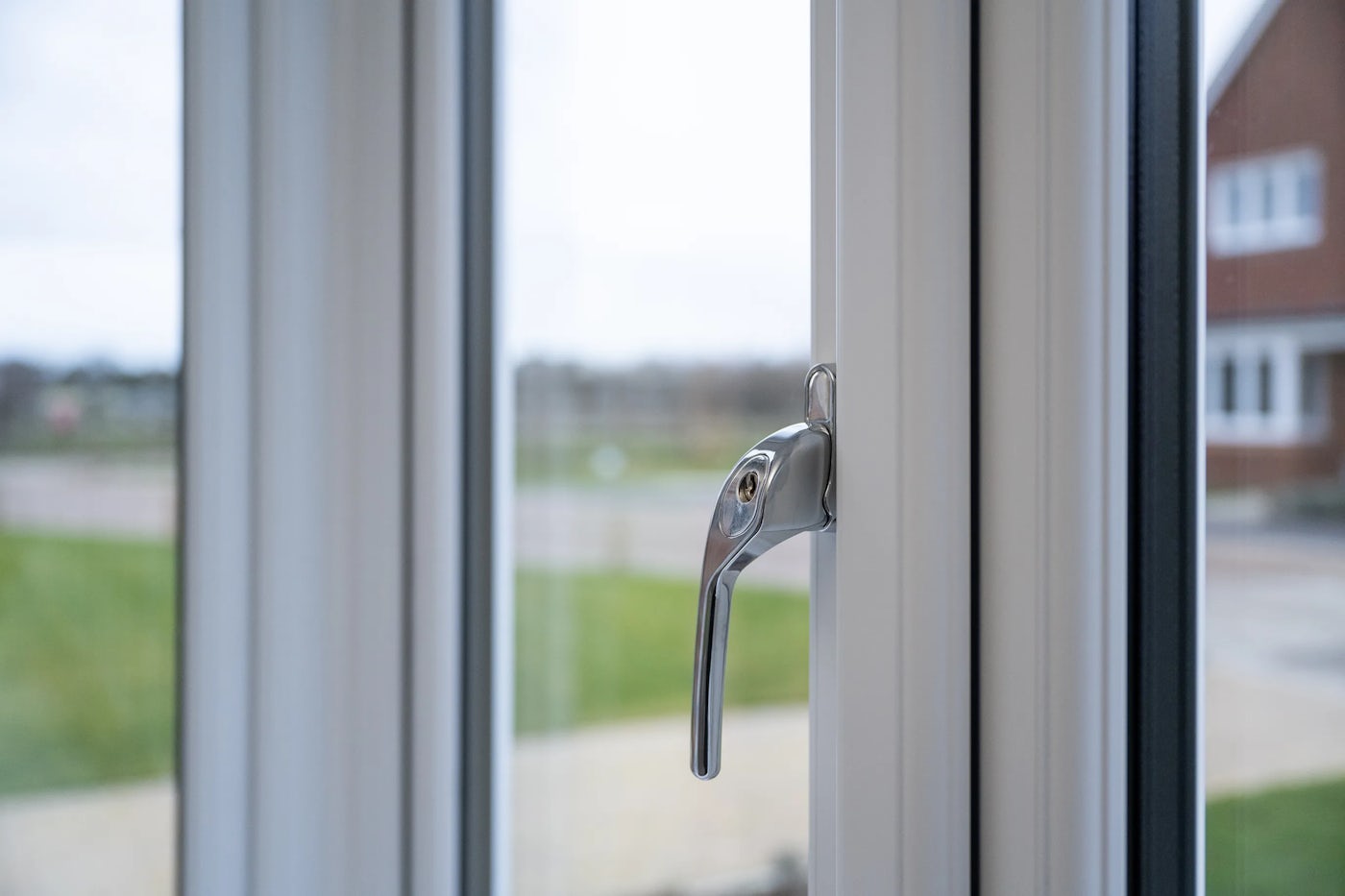 Close-up of a white-framed window featuring a sleek metallic handle with an integrated keyhole, indicating lock functionality. The handle is angled diagonally across the lower section of the frame, with natural light enhancing its smooth, reflective surface. In the background, a soft-focus view reveals green grass and a red brick building with white-framed windows, suggesting a secure and pleasant suburban setting