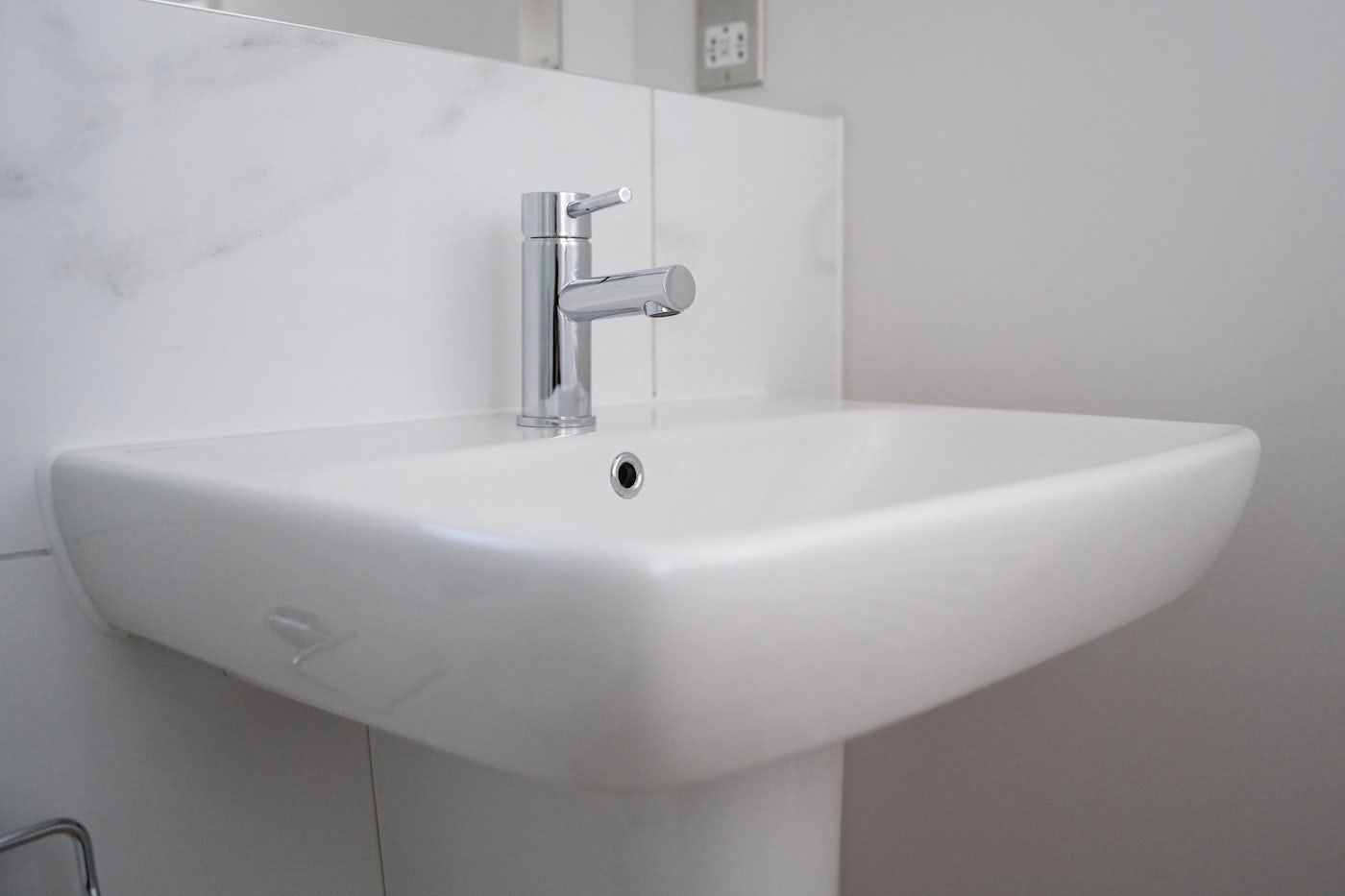 Close-up of a wall-mounted white ceramic sink with a clean, minimalist design, featuring a single-handle chrome faucet positioned centrally above the basin. The sink is set against a white wall with soft marble-like veining, adding subtle texture. An adjacent electrical outlet reinforces the domestic setting, while the overall scene is lit by natural light, enhancing the modern and practical feel of the space