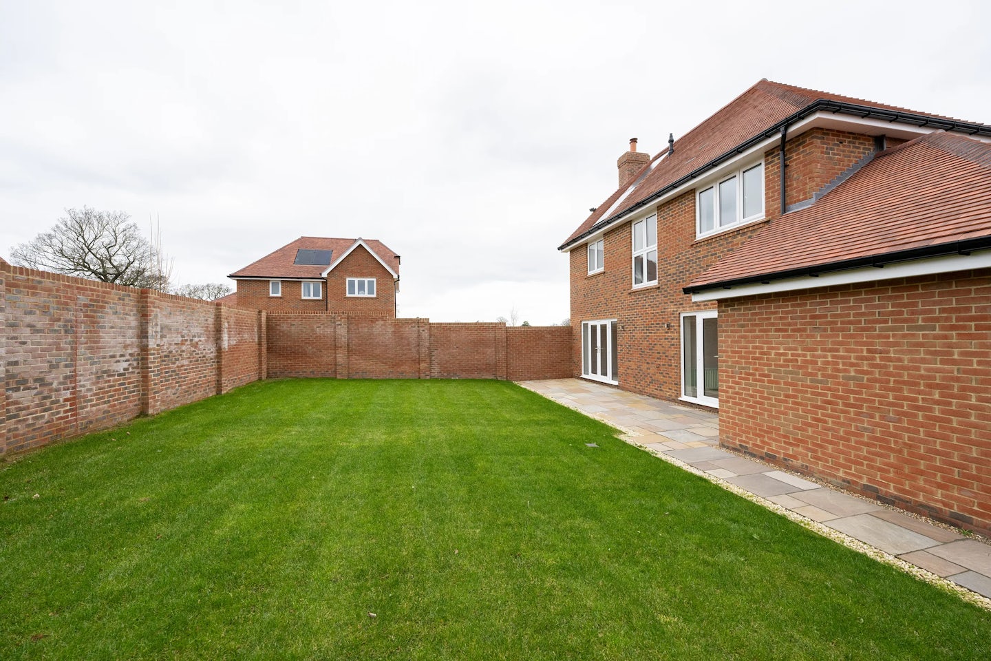 Suburban backyard featuring a neatly trimmed green lawn bordered by a low brick wall. The red-brick house with a red-tiled roof opens onto a paved patio area through large glass doors, offering an easy indoor-outdoor flow. Beyond the wall, another house and tall trees are visible beneath a grey, overcast sky, adding quiet depth to the private, enclosed setting.