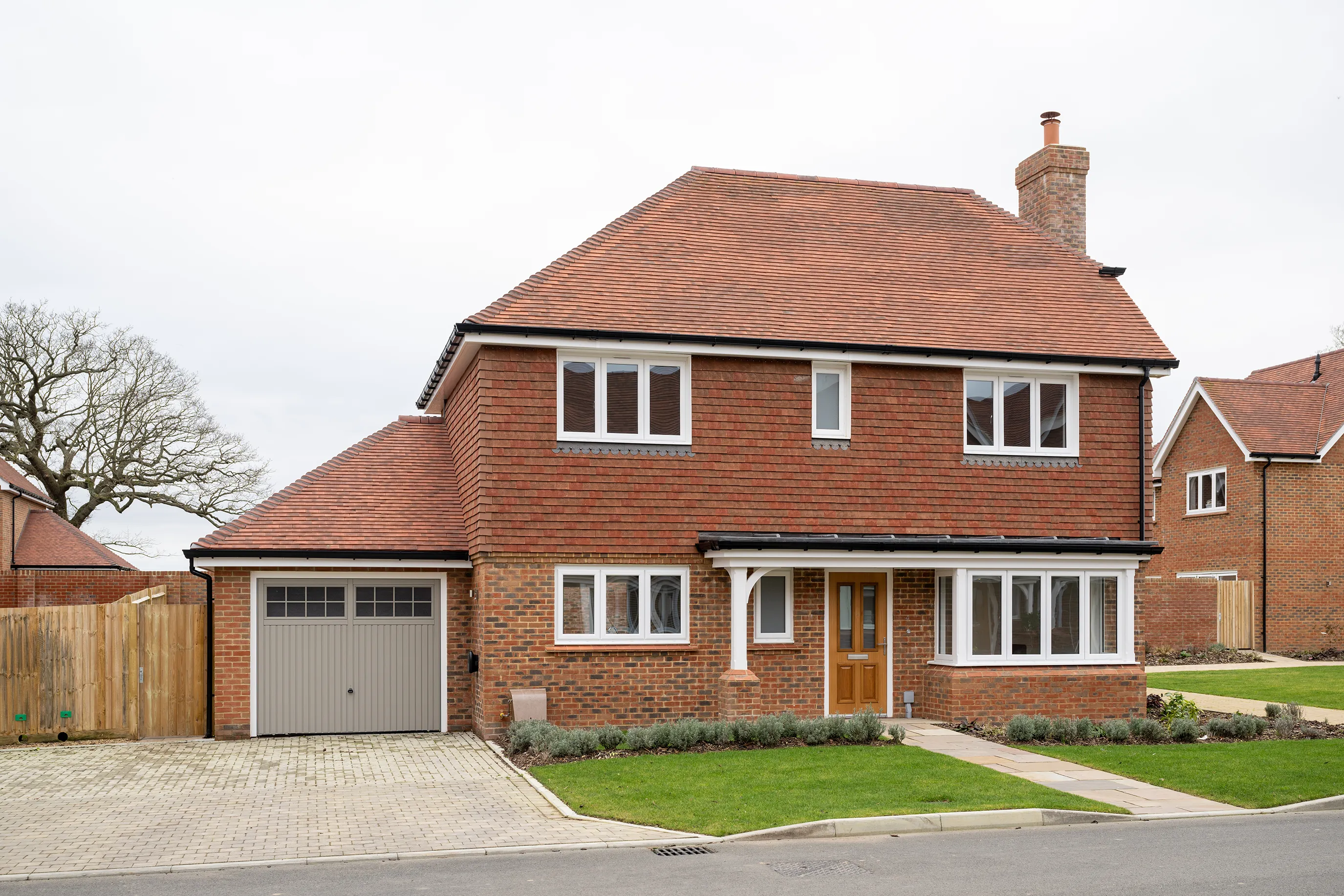 Modern two-story detached home with a red-tiled pitched roof and warm-toned brick exterior. Features include a grey garage door to the left, a wooden front door with glass panels, and white-framed windows arranged symmetrically across both floors. A neatly maintained front garden contains a small lawn and paved driveway, bordered by low hedges and planting. Another similarly styled house is visible nearby, and soft daylight under a partly cloudy sky contributes to the suburban calm and tidy architectural appeal