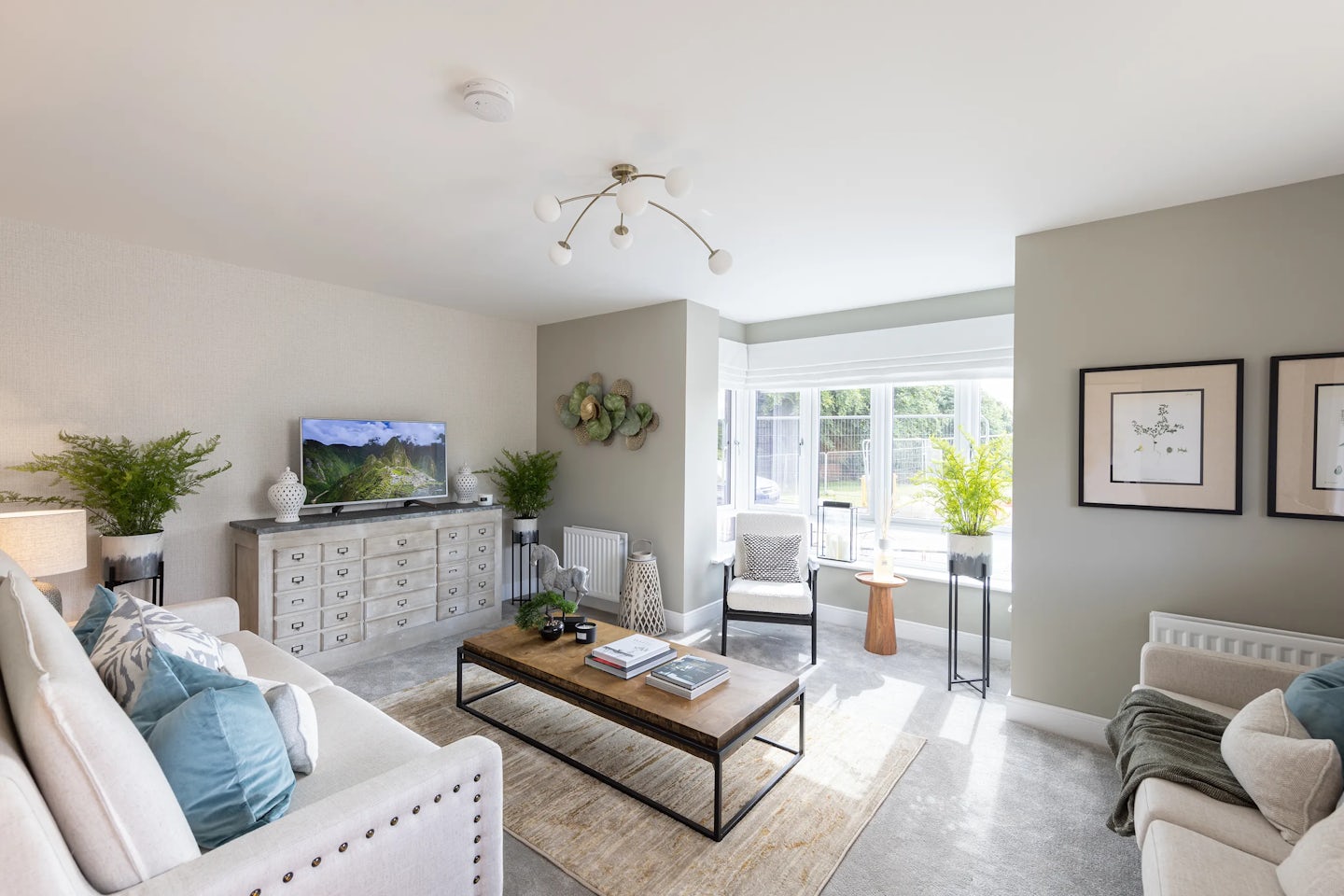 Bright, contemporary living room styled with a soft, neutral palette. A white sofa with blue and grey cushions faces a wooden coffee table adorned with books and decorative items. A sleek grey armchair and metal-framed side table with a potted plant flank the space, while a long grey sideboard supports a mounted TV displaying a serene nature scene. Large windows fill the room with daylight, and framed artwork adds character to the walls.