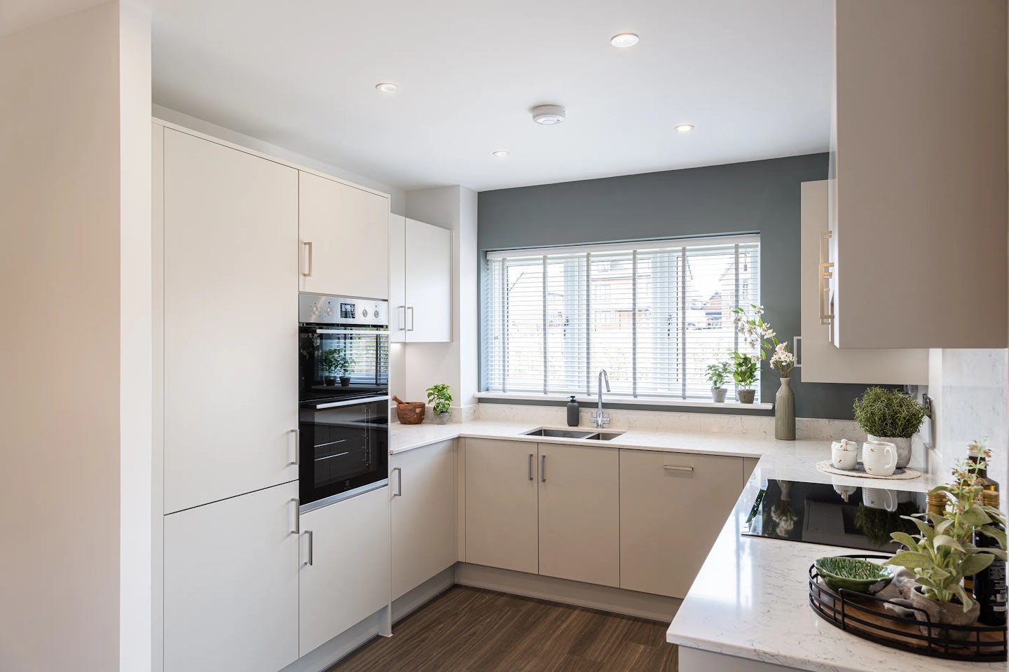 Contemporary kitchen with white cabinetry and a matching countertop, featuring built-in appliances including an oven, microwave, and sleek black-glass electric stovetop. A white sink sits beneath a wide window with horizontal blinds, allowing soft natural light to brighten the space. Green potted plants are placed strategically along the countertop and windowsill, infusing warmth and a biophilic touch. Recessed ceiling lights and light wood flooring complete the clean, minimal design