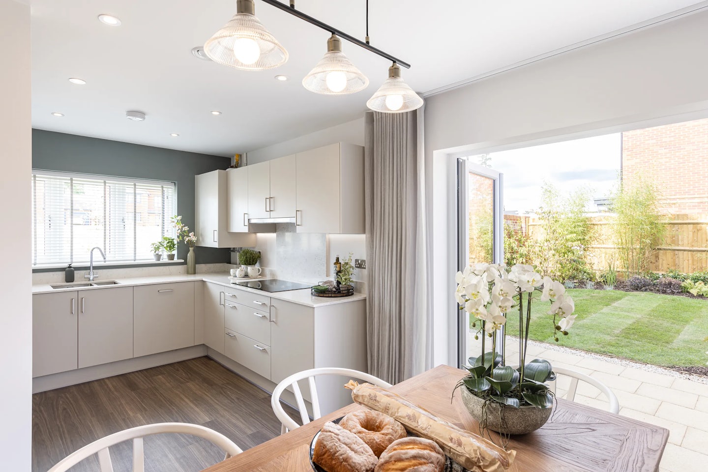 Open-plan kitchen and dining area with crisp white cabinetry, integrated stainless steel appliances, and a pale grey backsplash. The sink sits below a wide window with horizontal blinds, offering natural light across the sleek white countertop. A wooden dining table anchors the space, paired with white chairs and topped with a bowl of orchids and a breadboard, adding organic texture. Overhead, a trio of black pendant lights provides visual symmetry. In the background, large glass doors frame a view of the lush backyard featuring green grass, low planting, and a wooden fence—all contributing to the bright, serene ambiance of the multifunctional living space