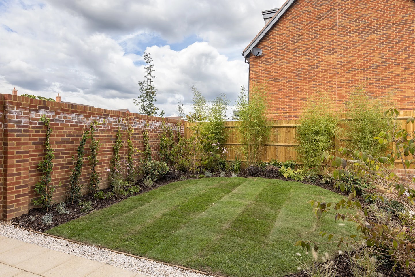 Well-groomed backyard garden with a neatly striped lawn framed by light paving stones and a strip of white gravel. Lush green climbing plants cover a brick wall on the left, while tall bamboo lines the wooden fence on the right. Mixed shrubs and foliage provide additional texture along the perimeter. In the background, a red-brick house with a pitched roof sits under a partly cloudy sky, creating a fresh, peaceful setting that emphasizes privacy and natural beauty