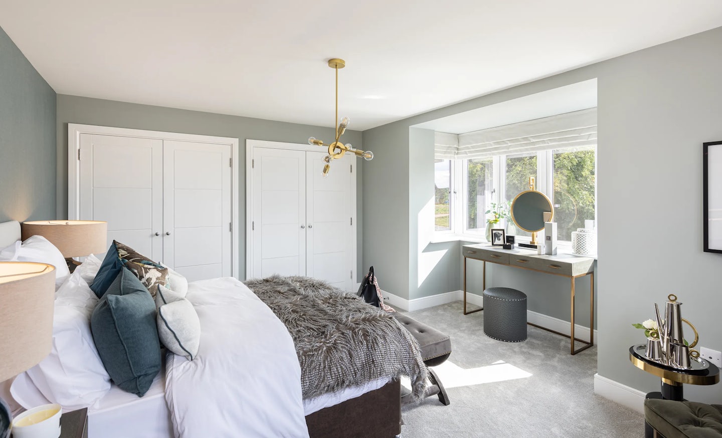 Stylish bedroom with a large double bed dressed in crisp white bedding, layered with teal and grey accent pillows and a textured grey throw. Twin bedside tables with coordinating lamps and accessories frame the bed, positioned against soft grey walls. To the right, a desk setup includes a round mirror, framed art, and decor arranged neatly beneath a window with white blinds. Below the window, a small stool and side table enhance the cozy layout, while a modern chandelier hangs above. Opposite the bed, double-door closets offer symmetry and storage. The light grey carpet and ambient lighting contribute to a serene, well-organized retreat