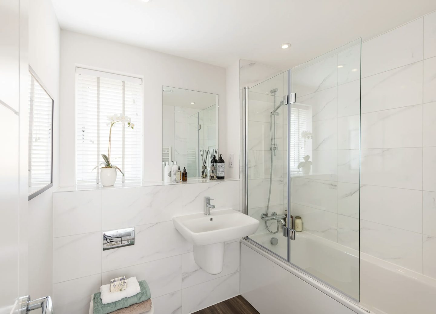 Bright, modern bathroom featuring a white sink with a chrome faucet mounted on a white tiled wall, complemented by a large mirror above. To the left, a small shelf displays bottles, a potted orchid, and other toiletries. A sleek bathtub with a glass shower screen and wall-mounted showerhead sits opposite two windows with white blinds, allowing soft natural light to fill the space. A compact stool beneath the sink holds neatly folded towels. The overall palette of white and light grey creates a clean, minimalist atmosphere with subtle elegance