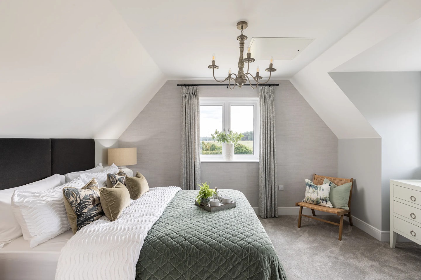Elegant bedroom with a sloped ceiling and large window framed by pale curtains, offering a view of lush greenery outside. A central double bed features white bedding with floral accent pillows, flanked by a side table holding a classic lamp. Above, a five-light chandelier adds sophistication, complementing the neutral-toned walls and soft flooring.