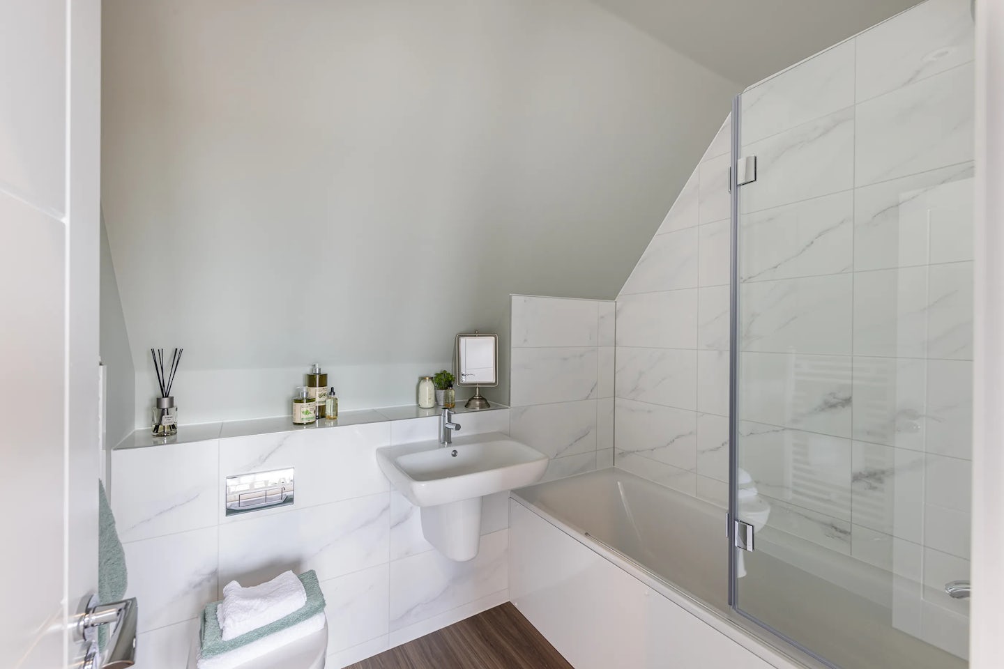 Compact, modern bathroom with white marble-effect wall tiles and a light wood-style floor. The space features a wall-mounted white ceramic sink beneath a small mirror shelf holding a soap dispenser, lotion bottle, and potted plant. To the right, a white bathtub with a glass shower screen is positioned under a sloped ceiling, while a neatly folded towel rests atop the adjacent toilet.