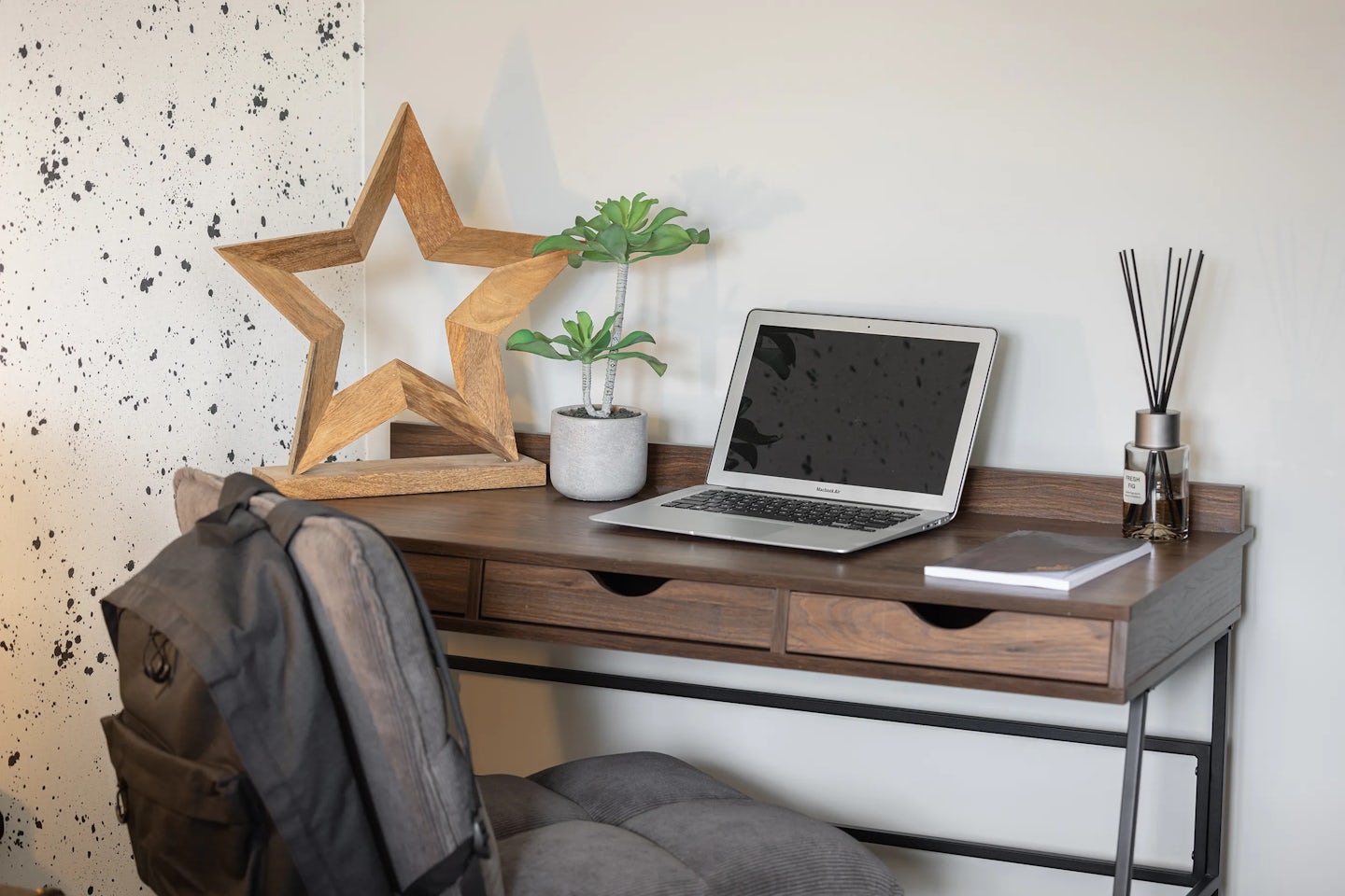 Minimalist workspace with a wooden desk featuring three drawers, topped by an open laptop, a notebook, a reed diffuser, a small potted plant, and a wooden star ornament. A grey cushioned chair sits in front of the desk, with a backpack hanging off its backrest. The backdrop includes a wall with a speckled pattern on the left, creating a subtly textured contrast.