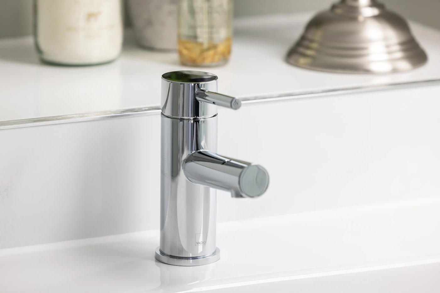 Close-up of a modern chrome faucet with a cylindrical base and single lever handle, mounted on a white countertop. The faucet’s reflective finish catches soft ambient light, highlighting its sleek, contemporary form. In the background, countertop accessories—including a glass jar with a white substance, another with a yellow substance, and a metallic lamp base
