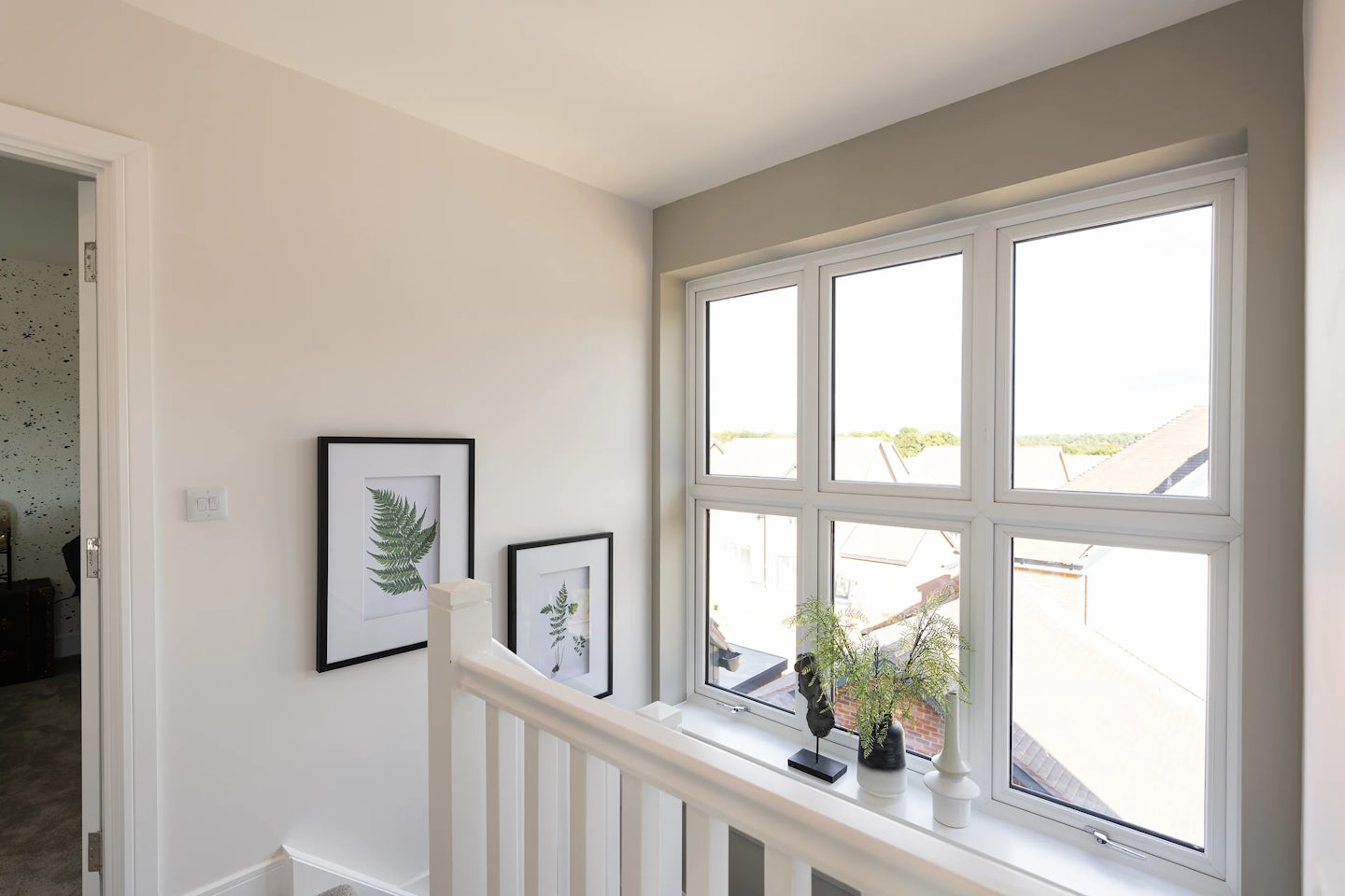 Bright and airy upper-floor landing featuring a large four-pane window that floods the space with natural light. Below the window, a shelf-like ledge displays two framed botanical prints, a potted plant, and a sculptural black ornament. The view through the window reveals distant rooftops under a clear sky. Light-toned walls and a white stair railing contribute to the calm, open atmosphere
