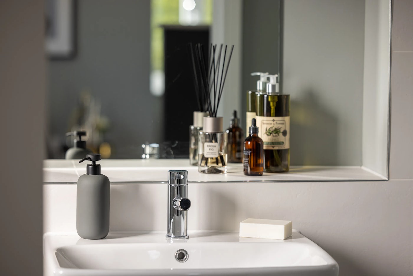 Bathroom sink area styled with a mix of modern functionality and personal care items. A white ceramic sink with a sleek chrome faucet anchors the foreground, accompanied by a grey soap dispenser on the left and a white soap bar on the right. Behind the sink, a mirror reflects an array of countertop accessories including a reed diffuser, a large pump bottle, and several smaller containers—one labeled “Fresh” and another featuring green botanical illustrations.