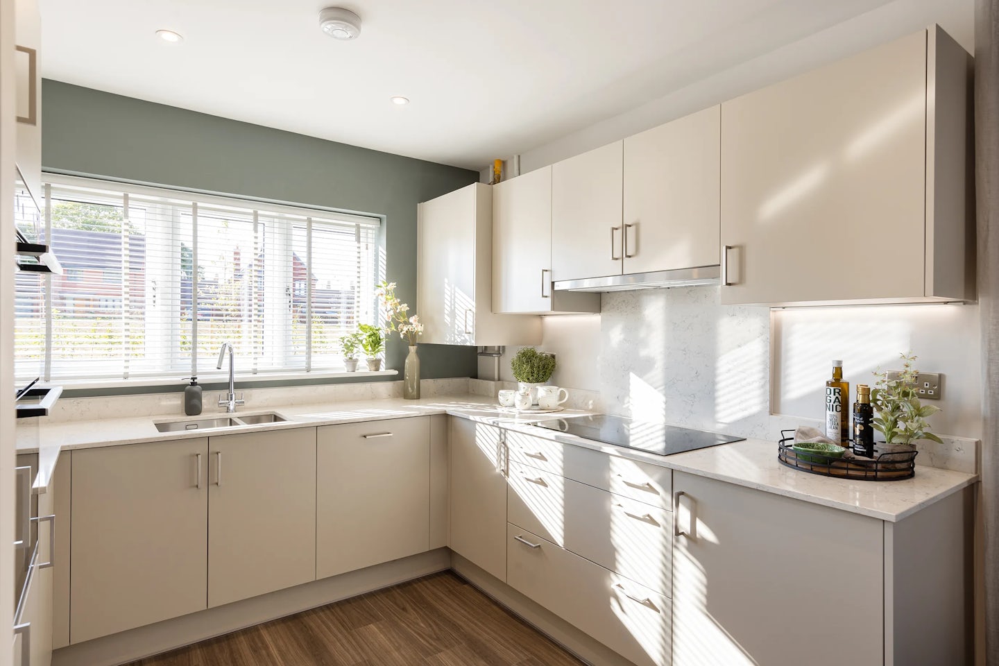 Bright contemporary kitchen with white cabinetry, matching countertops, and a light wood floor. A wide window with horizontal blinds sits above the sink, inviting natural light across the work surface. Decorative touches include a small potted plant by the faucet, a vase of tulips, and bottles of oil and vinegar arranged neatly along the right counter edge. A stainless steel stovetop with a sleek hood adds functionality