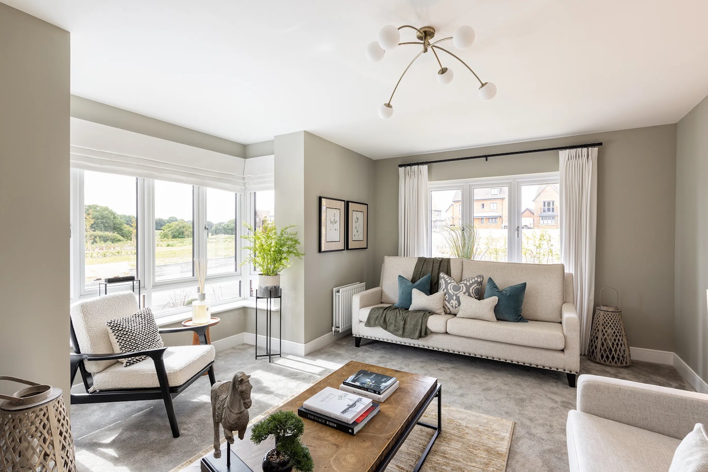 Tastefully styled living room with a large beige sofa adorned with blue and grey cushions, paired with a sleek black-framed grey armchair. A central wooden coffee table with a metal base holds books, a potted plant, and small decorative objects. Neutral-toned walls feature framed artwork, while white curtains frame tall windows that pour natural light into the space. A small side table with a lamp and plant sits near the window, and a modern multi-bulb ceiling fixture adds a contemporary touch. The soft grey carpet and balanced textures contribute to a cozy yet refined atmosphere