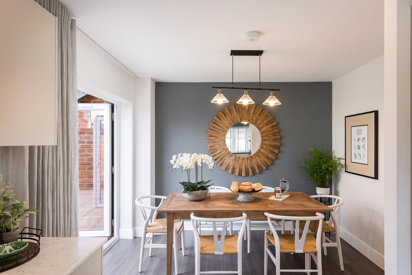 Stylish dining room featuring a rectangular wooden table surrounded by six white chairs with woven seats. Centerpiece decor includes a blooming orchid in a ceramic pot, a leafy plant, a tray of pastries, and a teapot—evoking hospitality and warmth. Above, a minimalist black light fixture with three pendant bulbs hangs over the table, complementing the clean-lined aesthetic. On the wall behind, a large round wooden-framed mirror and a framed art print add subtle character. To the right, a tall potted plant anchors the corner. A sliding glass door opens to the outdoors on the left, while part of a kitchen counter peeks in, complete with a basket and greenery.