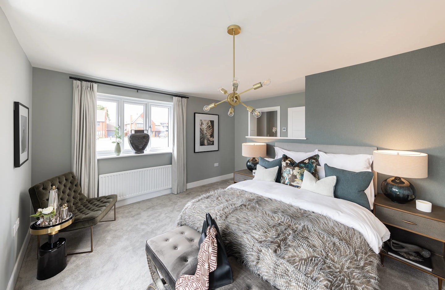 Elegant bedroom with a soft grey and white palette, centered around a plush double bed adorned with multiple accent pillows and a faux fur throw. Twin bedside tables support matching lamps and decorative accessories, while a distinctive multi-bulb chandelier hangs above. To the right, pale grey curtains frame a large window above a radiator, adding natural light to the serene setting. A green velvet armchair and a round side table with a tea tray sit beside the window, evoking comfort and ritual.