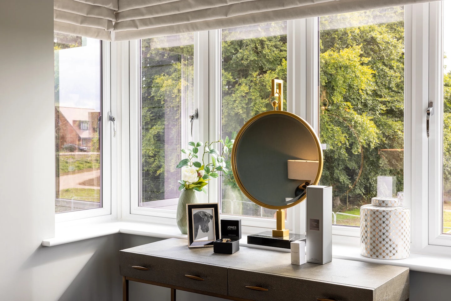 Bedroom vanity corner set beside a large window with a beige Roman shade, offering a serene view of lush greenery. A pale-toned table with gold-handled drawers serves as a surface for personal items: a gold-framed round mirror, a framed photo of a dog, a decorative jar with geometric patterns, a vase of green foliage, and two product boxes, one labeled “EVE LOM.” Soft daylight highlights the clean textures and muted palette, suggesting a tranquil space for reflection and daily ritual